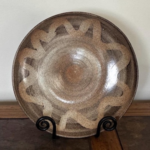 May include: A handcrafted ceramic bowl in shades of brown and beige. The bowl has a spiral pattern around the edge. It is presented on a black metal stand, highlighting its unique, artistic design. The bowl's diameter is approximately 25 cm.