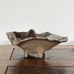 May include: A handcrafted ceramic bowl with a unique, organic shape. The bowl features a light tan base and a dark brown and grey rim. It is supported by three small feet and has a textured surface.