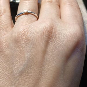Peut inclure: Une bague en argent avec une petite pierre précieuse transparente est portée au doigt. La bague a un anneau fin et une seule pierre surélevée. La main est mise au point, avec la bague bien en évidence. L'arrière-plan est flou.