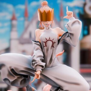 May include: A detailed figurine of a character in a dynamic pose, wearing a white outfit with a sun emblem and a gold crown. The figure is perched on a gray rock with orange accents, set against a blurred cityscape.