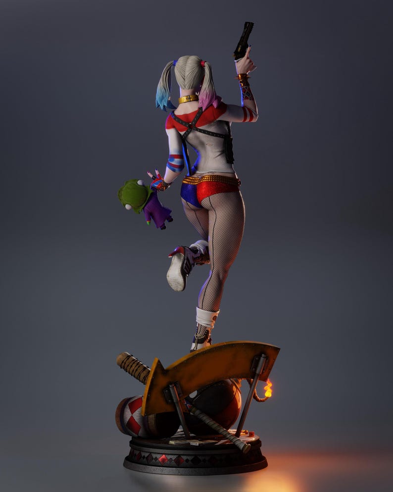 May include: A detailed figurine of a character with a white, red, and blue costume, holding a gun and a plush toy. The figure stands on a base with a mallet, a bomb, and a large yellow blade. The character has blonde pigtails with blue and pink tips.