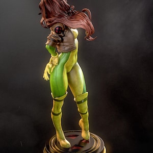 May include: A detailed figurine of Rogue from the X-Men. The statue features a green and yellow bodysuit, a brown jacket with an X-Men logo, and long auburn hair. The figure stands on a round base with a red X symbol.
