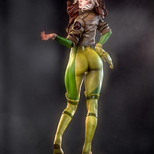 May include: A detailed figurine of a female character in a brown jacket, green gloves, and yellow and green jumpsuit. The figure has brown and white hair and is posed with one arm extended. The figure is standing on a black base.