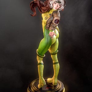 May include: A detailed figurine of a female character in a yellow and green bodysuit, brown jacket, and white-streaked brown hair. The figure is posed on a round base with a gold trim, showcasing a dynamic stance. The character's costume features a distinctive X-Men logo.