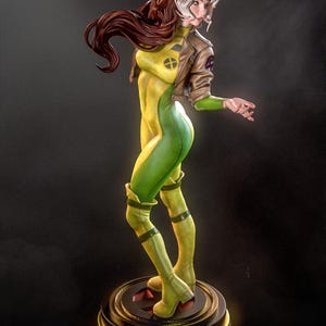 May include: A detailed figurine of Rogue from the X-Men. The statue features Rogue in a dynamic pose, wearing a yellow and green bodysuit, a brown jacket, and boots. Her hair is a mix of brown and white. The figurine is on a round base.