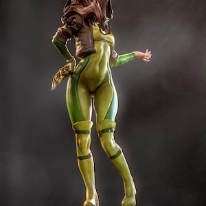 May include: A detailed figurine of a character in a green and yellow bodysuit, brown jacket, and brown hair. The figure is posed on a black base, with one arm extended. The figure has a dynamic pose, with a focus on the character's form and costume.