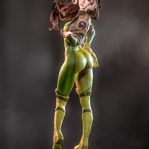 May include: A detailed figurine of a female character in a green and yellow jumpsuit, brown jacket, and boots. The figure has brown and white hair and is posed with one arm extended. The base is black and circular. The overall theme is comic book or superhero.