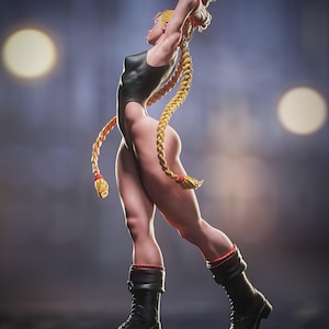 May include: A figurine of a woman in a black bodysuit, black boots, and long blonde braids. The figure is posed with arms raised, showcasing a muscular physique. Red accents are visible on the boots. The background is a blurred gradient of blue and gray with two bright circular lights.