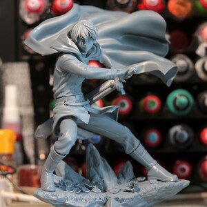 May include: A gray figurine of a person in a dynamic pose, holding a sword. The figure is wearing a cloak and boots, standing on a rocky base. The background is blurred, with various paint bottles visible.