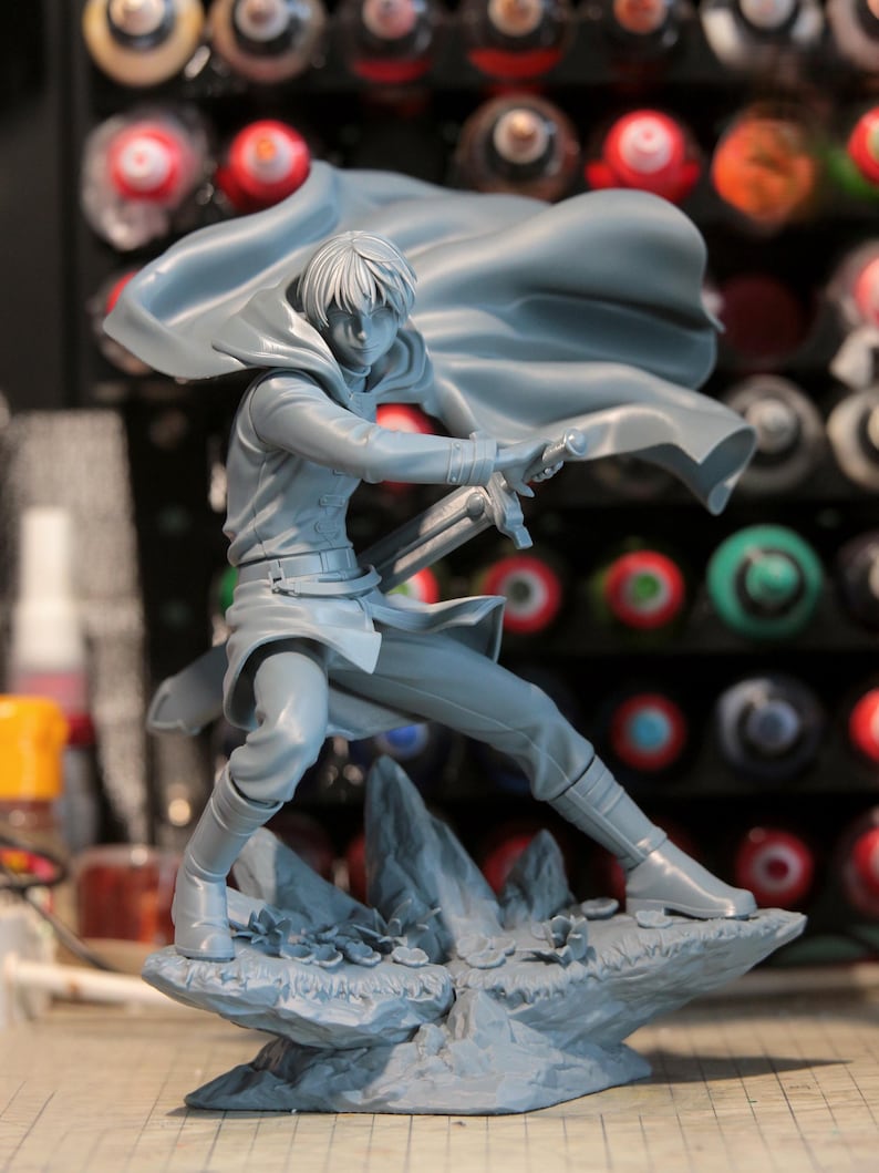 May include: A gray figurine of a person in a dynamic pose, holding a sword. The figure is wearing a cloak and boots, standing on a rocky base. The background is blurred, with various paint bottles visible.