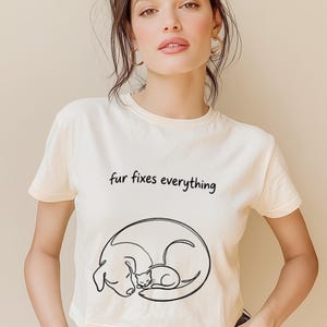 May include: Cream-colored crop top with the phrase "fur fixes everything" in black script. Below the text is a black line drawing of a dog and cat curled up together. The shirt is short-sleeved.