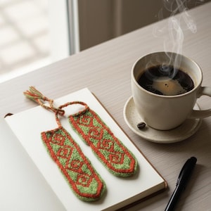 Puede incluir: Dos marcadores de ganchillo verdes y naranjas con un diseño geométrico descansan sobre un cuaderno abierto. Una taza de café con vapor ascendente se encuentra sobre un platillo. También hay un bolígrafo negro sobre la mesa.
