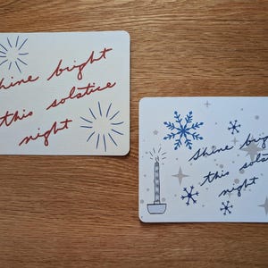 May include: Two rectangular cards with the text "Shine bright solstice this night". One card has red text and blue starbursts. The other card has blue snowflakes, stars, and a candle with gray text.
