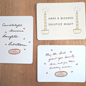 May include: Three white cards with handwritten text and illustrations. One card reads "Candlelight, Incense, Laughter, Libations." Another says "Have a Blessed Solstice Night." The third card reads "May the Gods grant you beauty, love, and joy this holiday season."