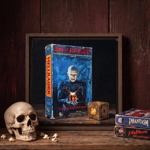 May include: A Hellraiser VHS tape box featuring a central figure with pins in their head, a gold-colored puzzle box, a human skull, and two additional VHS tapes labeled Phantasm and A Nightmare on Elm Street, all arranged on a wooden surface.