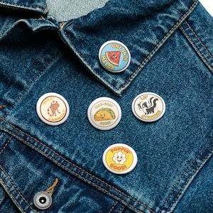 May include: A collection of round, colorful enamel pins with various cartoon designs and text. The pins include a watermelon slice, a taco, a skunk, a frog, and a popcorn graphic. The pins are attached to a blue denim jacket.