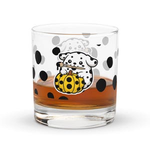 May include: A clear glass tumbler featuring a cartoon Dalmatian dog painting a yellow pumpkin with black spots. The glass has black and grey polka dots and is filled with a brown liquid.