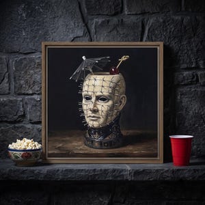 May include: A framed square painting depicting a beige, head-shaped ceramic mug covered in silver pins, resembling a character with black eyes. The mug features a black collar base, a small black cocktail umbrella, and a single red cherry on a pick.