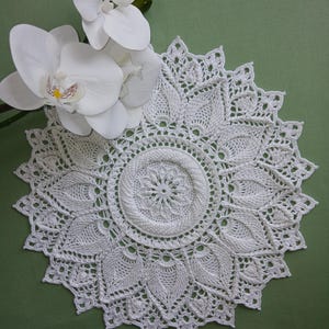 May include: A round white crochet doily featuring an intricate floral-inspired pattern with a central circular motif and radiating leaf-like segments. The doily has a scalloped edge and is displayed alongside a white orchid flower with green buds.
