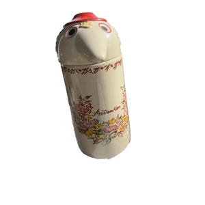 Puede incluir: Un tarro de cerámica vintage con cara de pájaro y sombrero rojo. El tarro cilíndrico presenta diseños florales y la palabra "Andenken". El tarro color crema tiene detalles florales rojos, amarillos y rosas.
