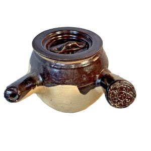 May include: A brown and tan ceramic vessel with a lid and two spouts. The vessel has a rounded body and a dark brown lid with a circular design. The spouts extend from the sides, one with a hole and the other with a decorative end.