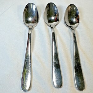 Puede incluir: Tres cucharas de acero inoxidable con cuencos ovalados y mangos largos y delgados. Las cucharas están dispuestas una al lado de la otra, reflejando la luz. Las cucharas son probablemente para comer o servir.