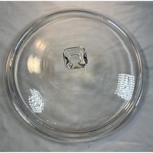 May include: A circular, clear glass lid featuring a smooth, slightly domed surface and a central, square-shaped knob handle. The lid has a reinforced outer rim designed to fit securely onto a corresponding container or cookware piece.