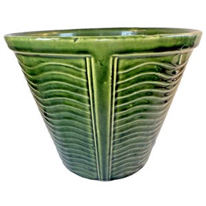Puede incluir: Una maceta de cerámica de color verde oscuro con acabado brillante, que presenta un borde ancho y acampanado y un cuerpo cónico. La superficie exterior está decorada con paneles verticales que contienen líneas horizontales onduladas en relieve.