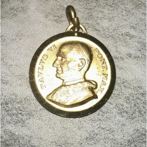 Pode incluir: Pingente dourado com o perfil de um homem e a inscrição "PAVLVS VI PONT. MAX". O pingente circular tem uma argola na parte superior para pendurar.