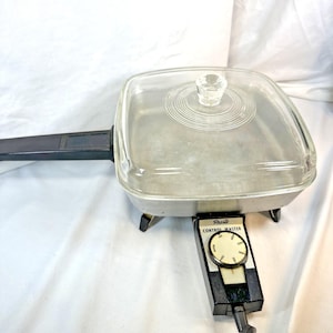 Puede incluir: Una sartén eléctrica cuadrada con acabado en aluminio plateado, mango negro y tapa de cristal transparente con pomo central. La base incluye un dial de temperatura negro Presto Control Master con marcas blancas y una aguja indicadora.