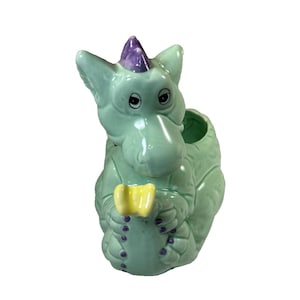 May include: A light green ceramic dragon figurine, holding a yellow object. The dragon features a purple horn and purple accents. The figurine has a glossy finish and is designed to hold small items. A decorative piece.
