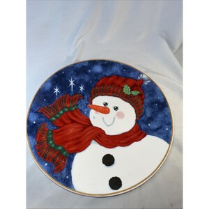 May include: A round decorative plate featuring a snowman design. The snowman has a carrot nose, black coal eyes and buttons, and a red scarf and hat. The plate's border is dark blue with white stars and a gold rim.