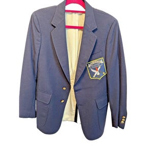 May include: A navy blue blazer with gold buttons and a Prep School Delinquent patch on the front. The blazer has a light-colored lining and is hanging on a pink hanger. The blazer has two front pockets.