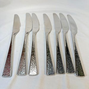 Puede incluir: Juego de ocho cuchillos de mantequilla de acero inoxidable. Cada cuchillo presenta un mango texturizado y una hoja curva. Los mangos tienen un diseño martillado y las hojas son lisas y brillantes. Los cuchillos están dispuestos sobre una superficie blanca.