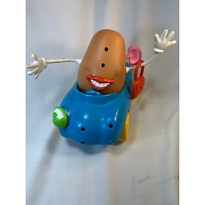 Könnte beinhalten: Ein skurriles Spielzeug mit einer kartoffelförmigen Figur mit einem lächelnden Mund, drei Augen und weißen Händen. Die Figur sitzt in einem blauen Auto mit gelben Rädern und einem grünen Scheinwerfer. Eine rote Handtasche hängt an der Seite.