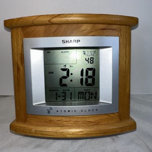 May include: A Sharp brand atomic clock with a light brown wood-grain frame and a silver-colored rectangular face. The digital display shows the time 2:18 AM, the date 1-31, the day MON, and a small map icon of the United States in the corner.