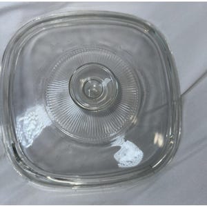 May include: A square, clear glass lid with rounded corners, featuring a central circular knob handle. The surface of the lid is decorated with a radial sunburst pattern of fine, embossed lines radiating outward from the center knob to the edges.