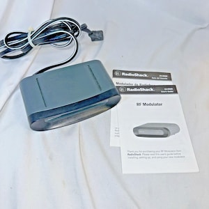 May include: A gray RadioShack RF Modulator with a black base and a connected cable. The image includes the user's guide and a small RadioShack label. The text on the guide reads "RF Modulator".