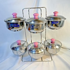 May include: A stainless steel spice rack with six small, round containers. Each container has a pink knob on the lid. The rack is made of silver metal and has a tiered design, with three containers on top and three below.