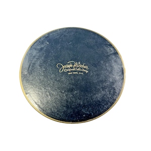 May include: A round, navy blue container with a gold rim. The text "Joseph Fleischer Original Custom Design NEW YORK, N.Y." is printed in gold script. The container has a textured surface.