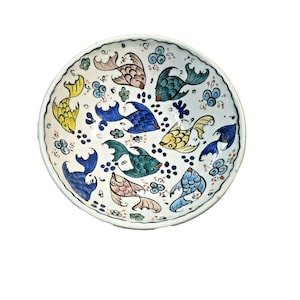 Puede incluir: Un cuenco redondo de cerámica blanca con un diseño pintado a mano de peces coloridos. Los peces son en tonos de azul, verde, amarillo y rosa, con detalles negros. El cuenco tiene un borde festoneado y está decorado con detalles florales.