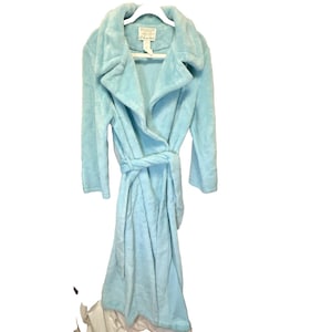 May include: A light blue plush robe featuring a wide collar, long sleeves, and a matching tie belt at the waist. The garment includes a white interior label at the neckline with visible text. The fabric has a soft, fuzzy texture throughout the design.
