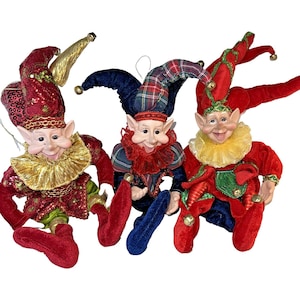 Puede incluir: Tres figuras de elfos caprichosos con caras sonrientes, orejas puntiagudas y atuendos festivos. Cada elfo lleva un sombrero y un cuello de bufón de diferente color, con detalles en rojo, verde y dorado. Los elfos posan con las piernas colgando, ideal para la decoración navideña.