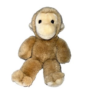 May include: A soft toy monkey in shades of brown and cream. The monkey has a light brown body, a cream-colored face, ears, and hands, and black eyes. It is sitting with its legs outstretched, and the fur appears soft.