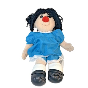 Puede incluir: Muñeca de peluche con cabello negro de hilo, nariz roja y rostro sonriente. La muñeca viste un vestido azul con ribete de encaje blanco, pantalones cortos a juego, calcetines blancos y zapatos negros. Juguete infantil.