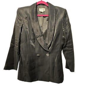 May include: A dark gray blazer with a satin finish, featuring a shawl collar and two decorative buttons. The blazer is hung on a pink hanger. The label reads "Cachet".