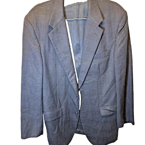 Può includere: Un blazer blu scuro con chiusura a due bottoni, revers a lancia e due tasche anteriori. La giacca è realizzata in un tessuto strutturato ed è appesa a una gruccia di metallo. La fodera interna è di una tonalità di blu più chiara.
