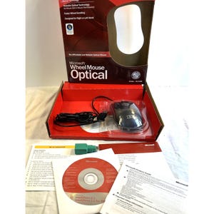 May include: A black Microsoft Wheel Mouse Optical with a cord, housed in its original red and white cardboard packaging. The set includes a green USB to PS/2 adapter, a red installation CD, and a printed Microsoft product guide document.