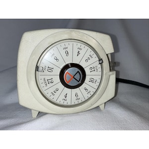 以下が含まれることがあります： 円形の文字盤が付いた、クリーム色のヴィンテージ目覚まし時計。文字盤には、AMとPMの表示とともに、放射状に時間が表示されています。時計は質感のある四角いボディで、黒い電源コードが付いています。