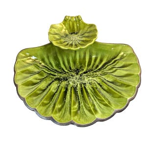 May include: A lime green ceramic serving dish shaped like a large scallop shell with a smaller, attached shell-shaped bowl at the top. The surface features a glossy glaze with deep ridges radiating from the center, transitioning to dark brown edges.